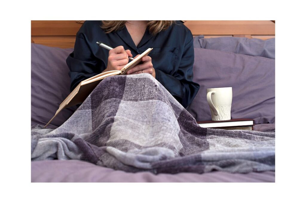 Woman writing in bed with covers over her legs and a coffee cup balanced on a book.