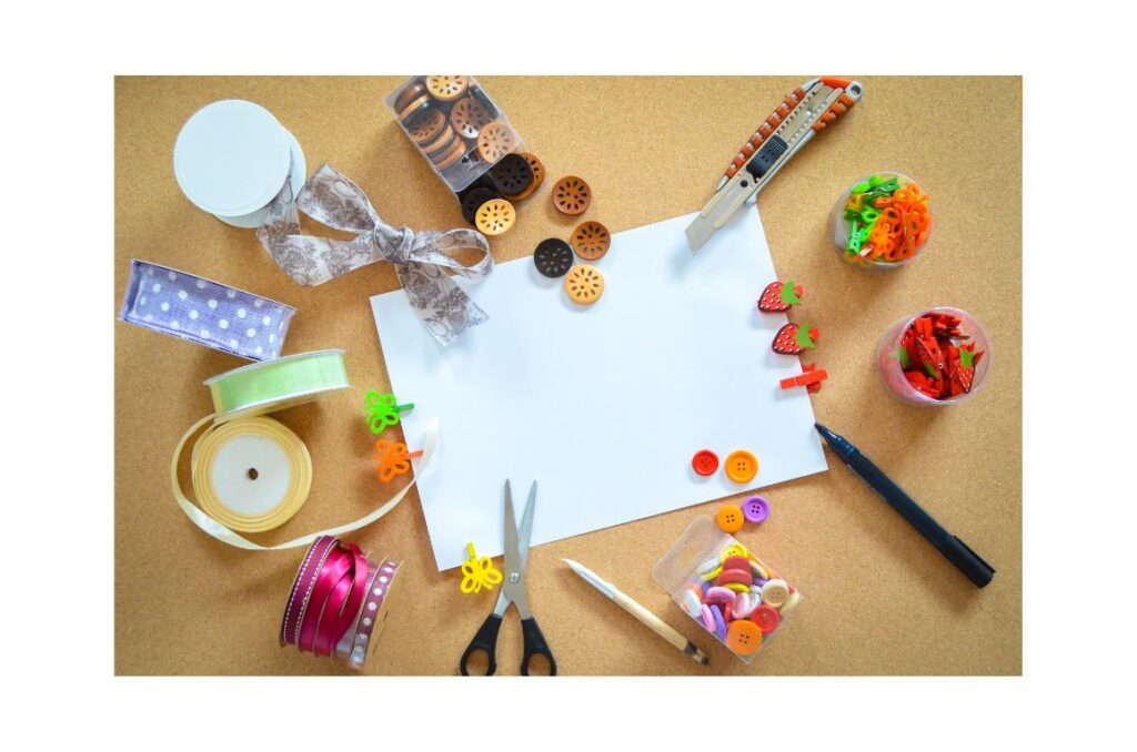 A variety of art supplies, such as ribbons, paints, scissors,etc. around a blank sheet of paper.